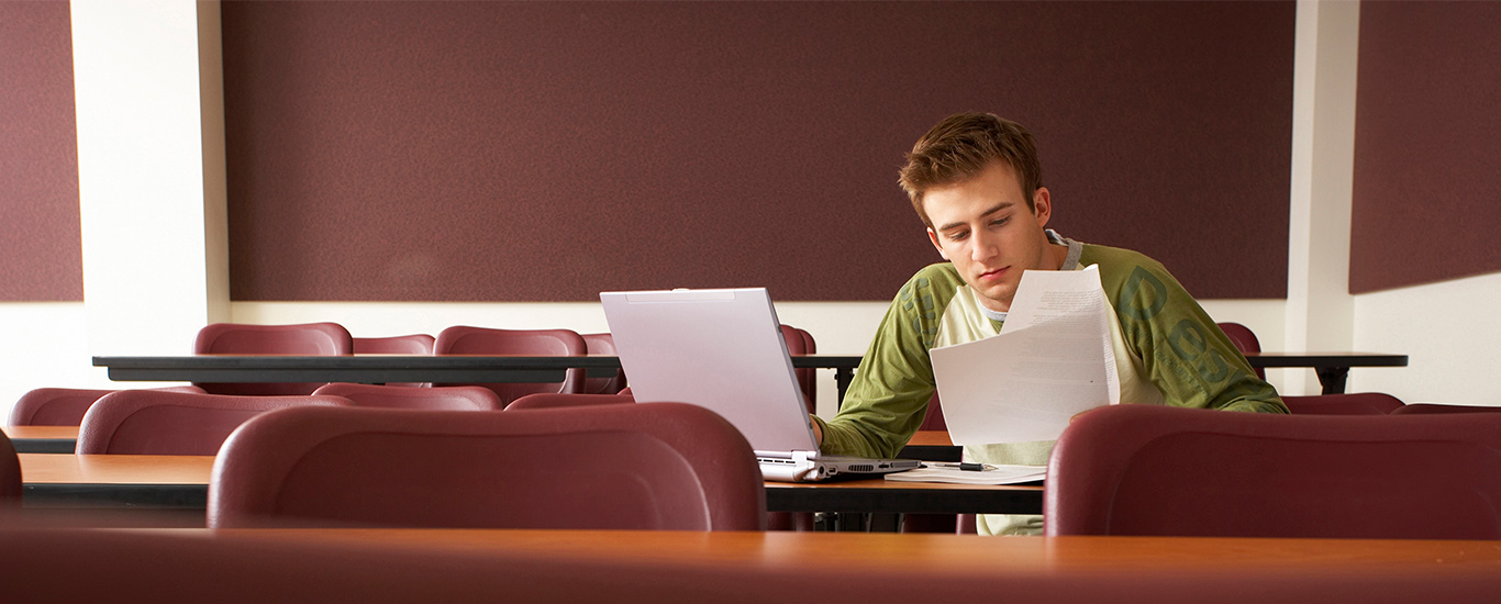 An image of a young man sitting alone in a lecture hall and looking over paperwork and at his laptop.